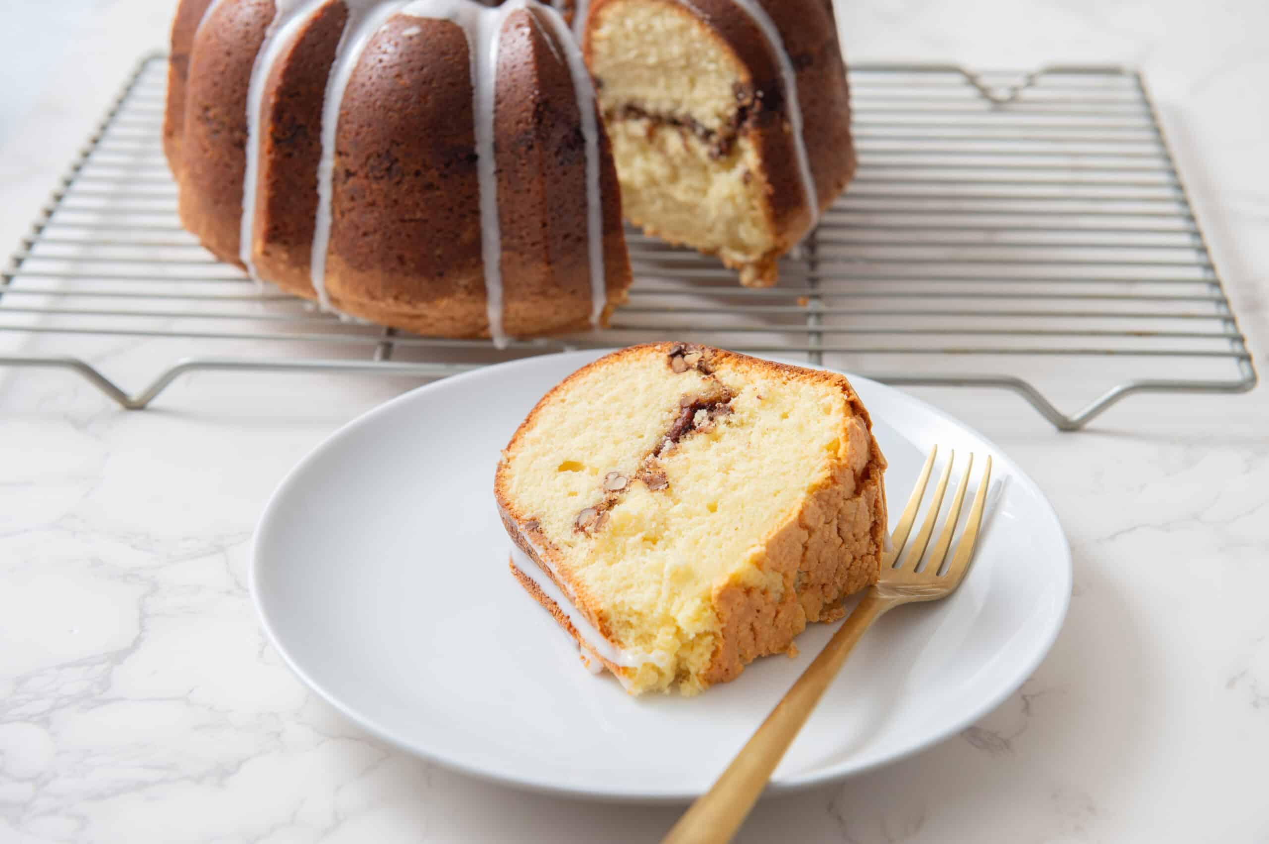 Fresh vanilla chiffon cake with chocolate chip filling, topped with white icing drizzle, on a white plate with a gold fork in front of a cooling rack. Perfect for dessert or tea time.