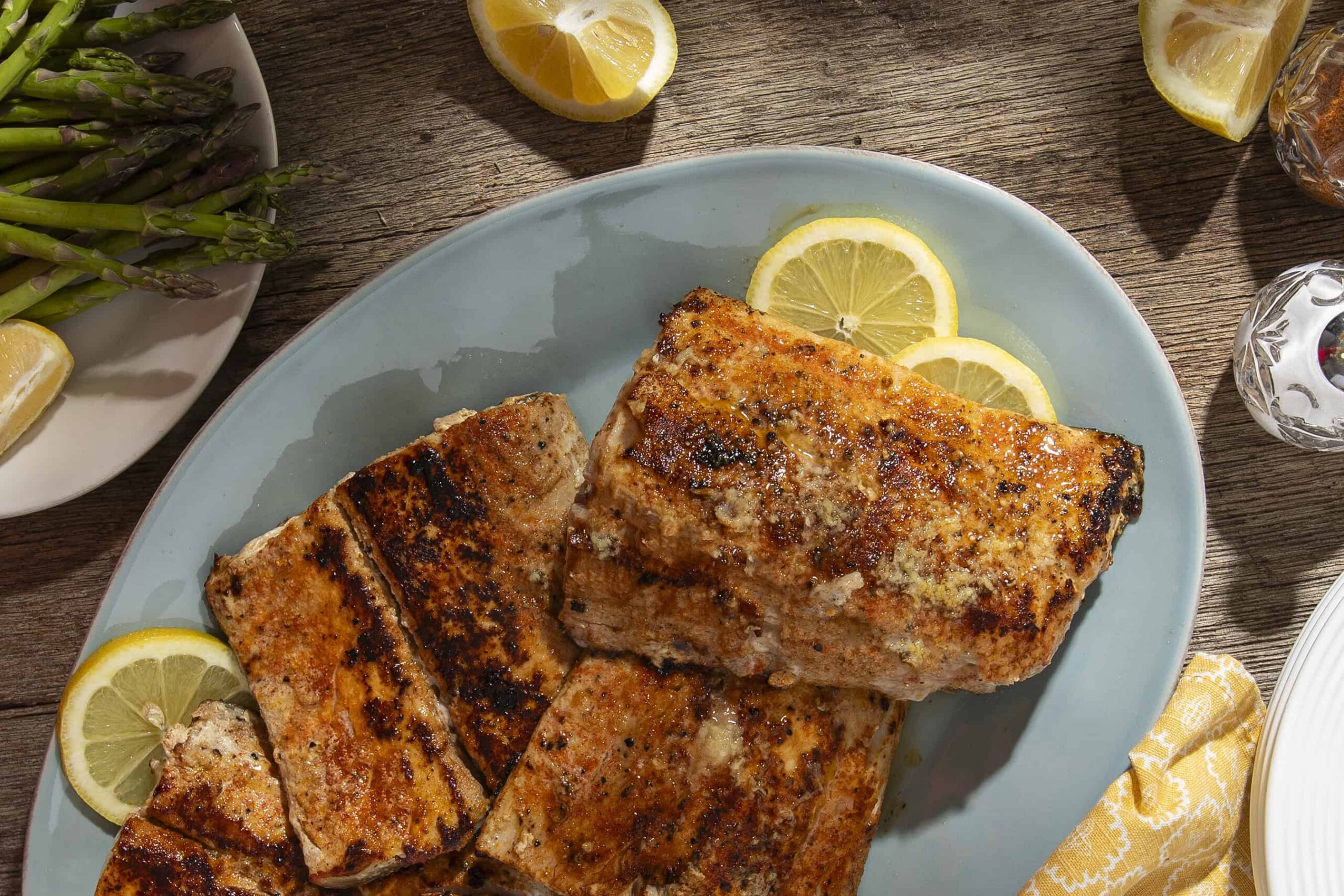 Succulent grilled salmon fillets garnished with lemon slices on a light blue plate, featuring a rustic wooden table setting with fresh asparagus and lemon wedges for healthy, delicious seafood meals.