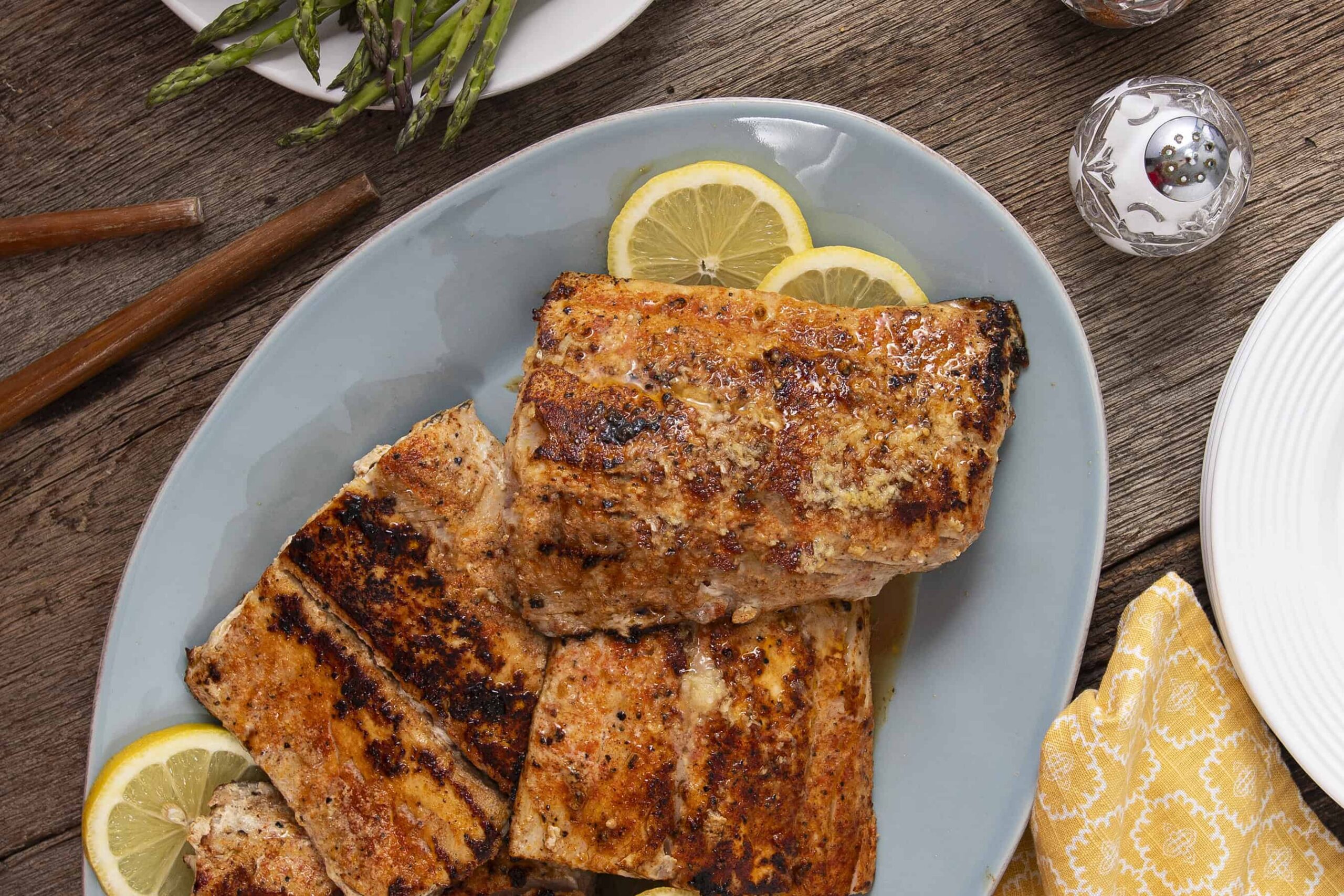 Juicy grilled salmon fillets garnished with lemon slices on a blue plate, served on a rustic wooden table with fresh asparagus and a yellow patterned napkin for a healthy seafood dinner.