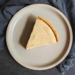 Creamy cheese cheesecake slice on a white plate with a graham cracker crust, placed on a dark textured background with a navy blue cloth. Perfect for dessert lovers and cheesecake recipes.