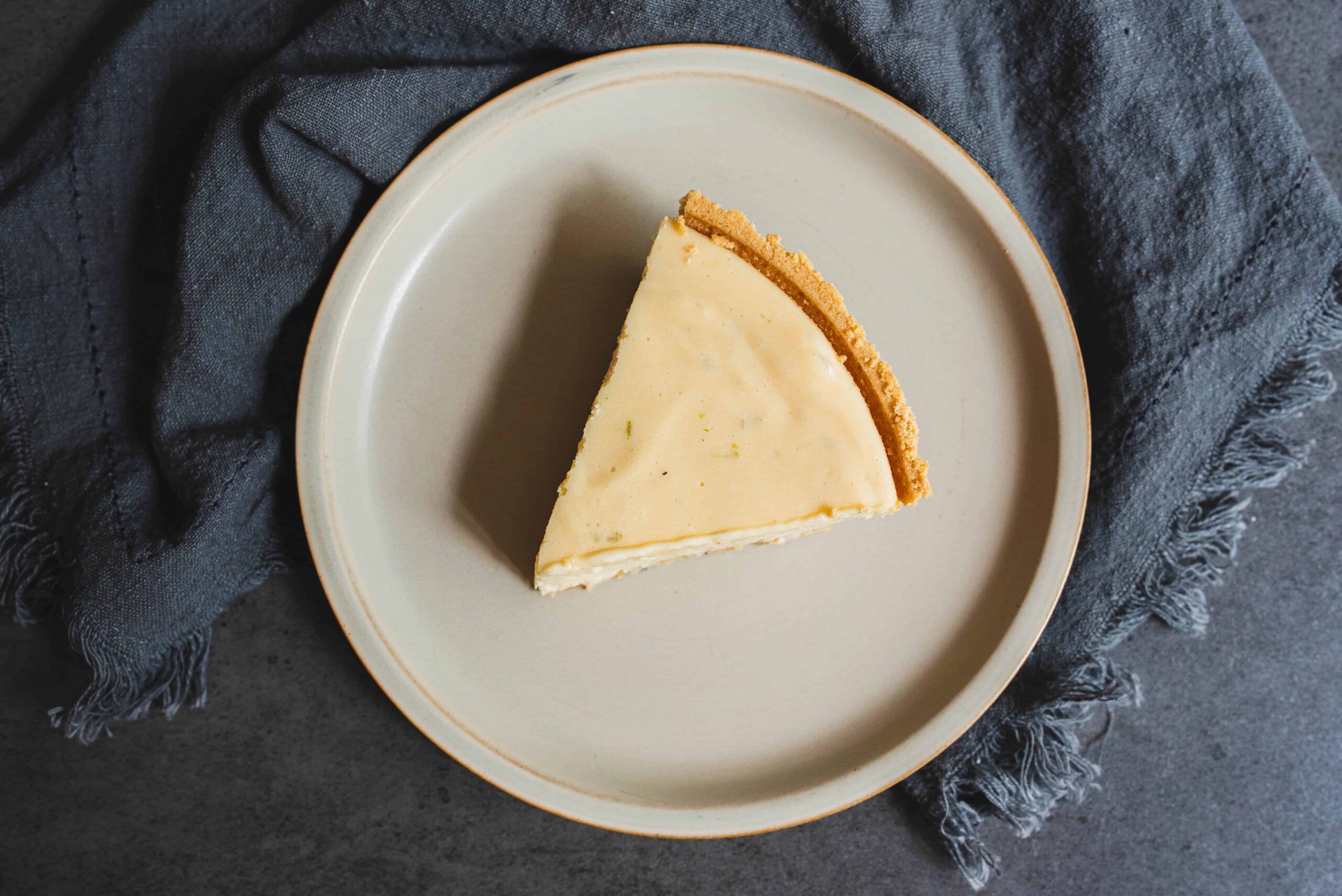 Creamy cheese cheesecake slice on a white plate with a graham cracker crust, placed on a dark textured background with a navy blue cloth. Perfect for dessert lovers and cheesecake recipes.