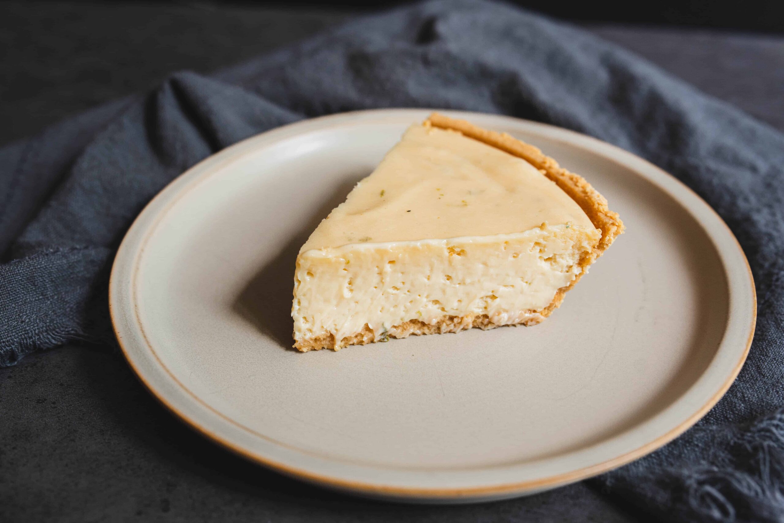 Creamy cheesecake slice on a neutral plate, with a graham cracker crust, perfect for dessert or special occasions, served on a dark textured background.