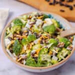 Fresh broccoli salad with black beans, shredded cheese, bacon bits, and chopped onions in a ceramic bowl on a light surface.