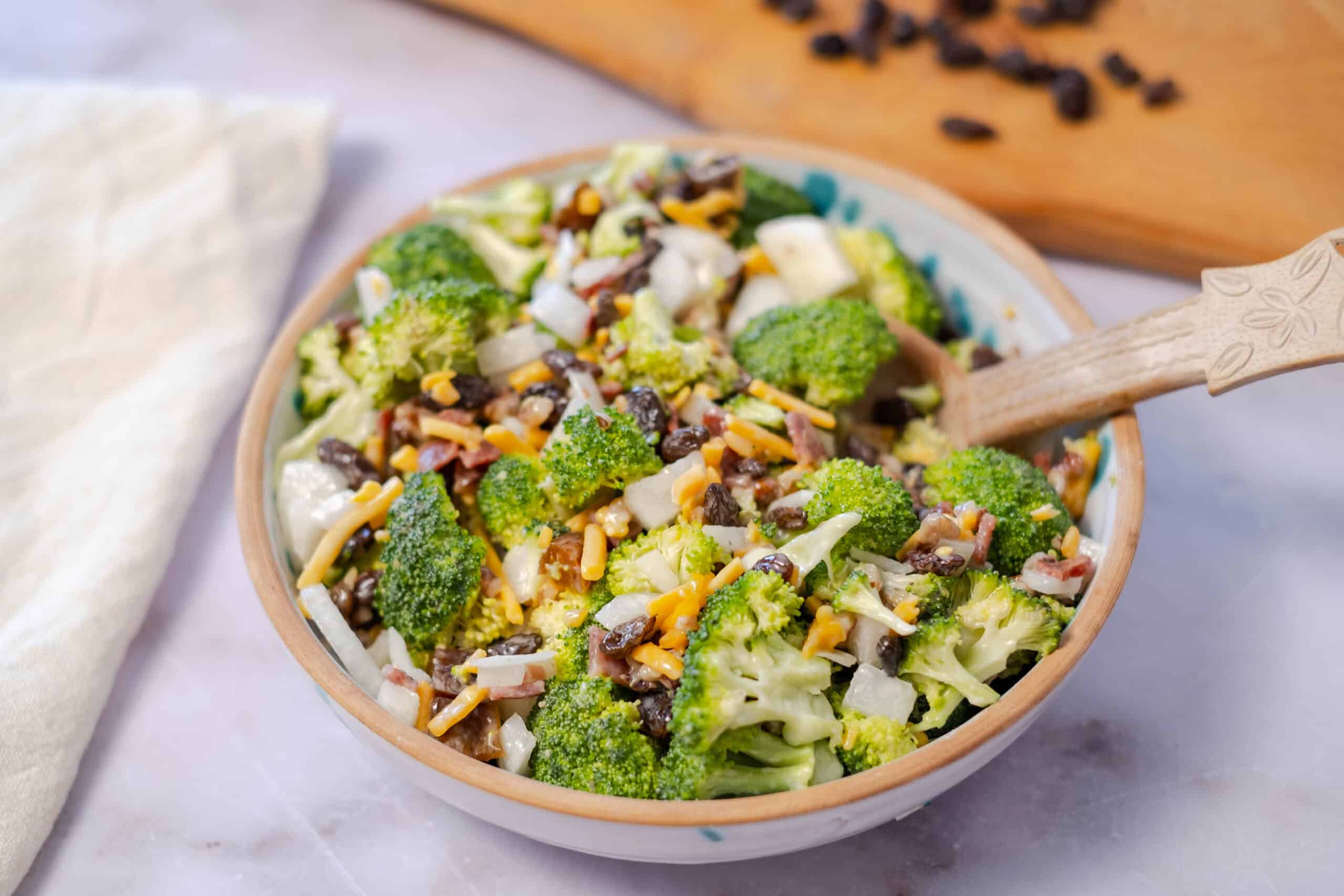 Fresh broccoli salad with black beans, shredded cheese, bacon bits, and chopped onions in a ceramic bowl on a light surface.