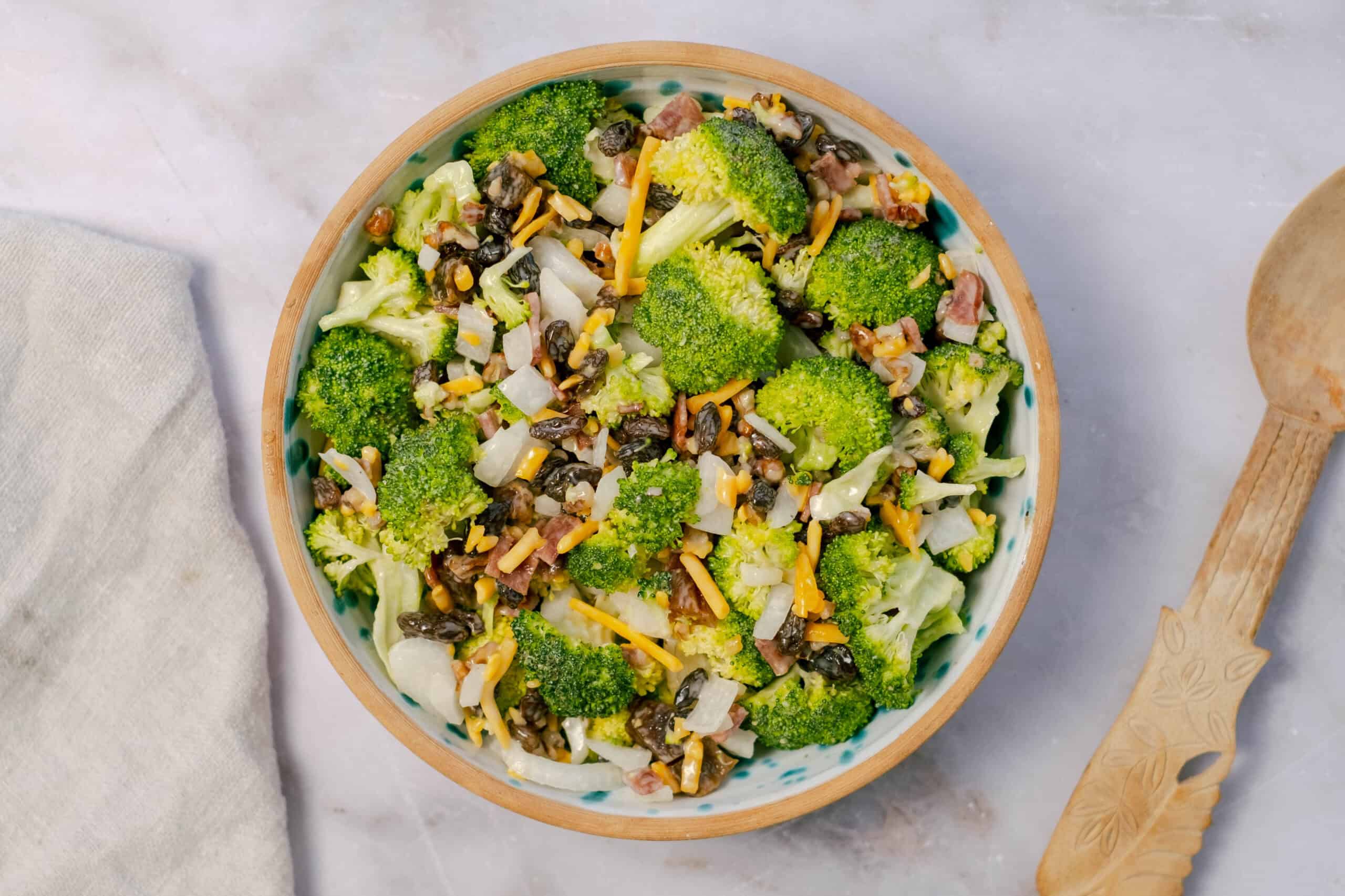 Fresh broccoli salad with shredded cheese, bacon bits, sunflower seeds, and chopped onions in a wooden bowl. Healthy and tasty vegetable side dish for any meal.