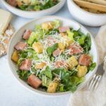 Crispy chicken Caesar salad with fresh greens, grated cheese, croutons, and bacon bits served in a white bowl on a light surface.