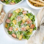 Fresh homemade Caesar salad with crispy croutons, shredded cheese, romaine lettuce, and ham, served in a white bowl with cheese, forks, and a wooden cutting board in the background.