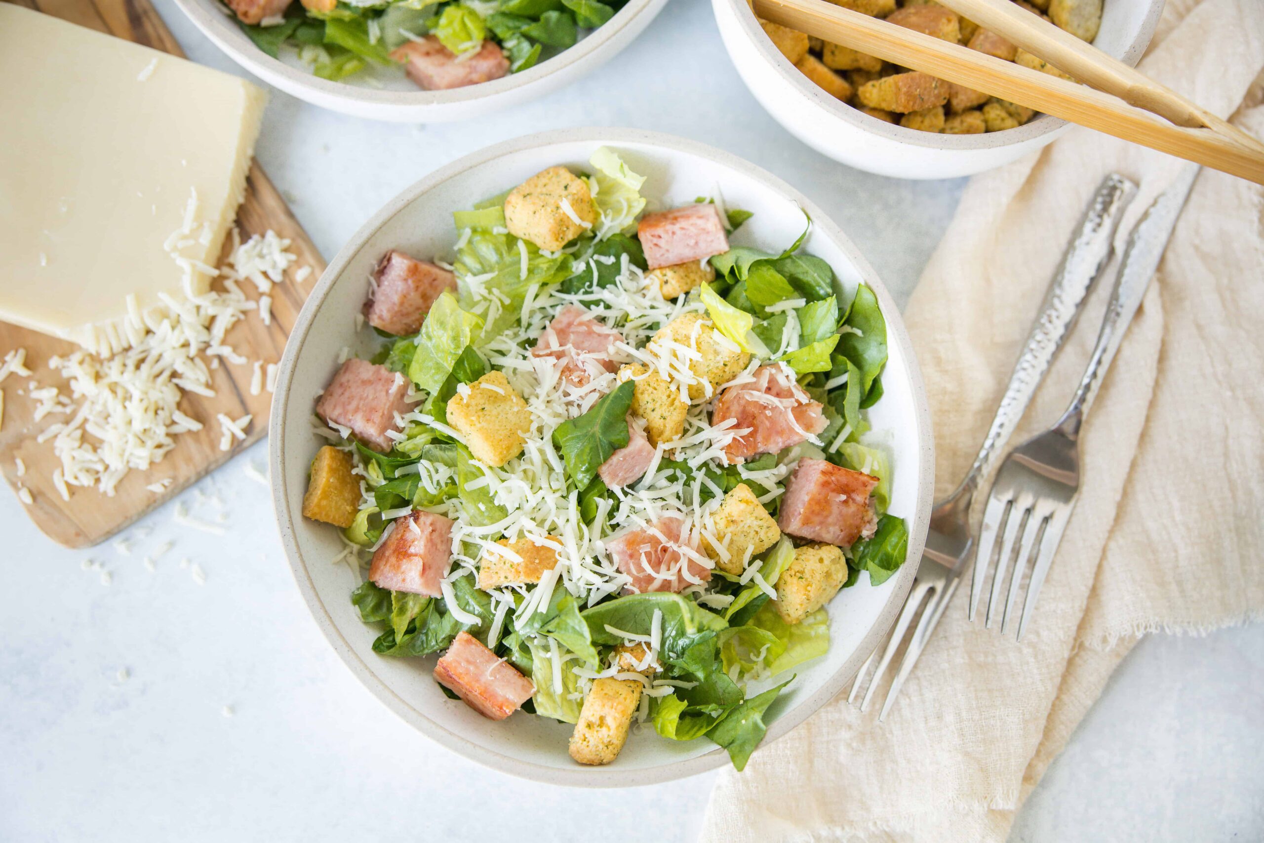 Fresh homemade Caesar salad with crispy croutons, shredded cheese, romaine lettuce, and ham, served in a white bowl with cheese, forks, and a wooden cutting board in the background.