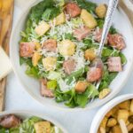 Fresh Caesar salad with crisp romaine lettuce, grilled chicken, shredded Parmesan cheese, and crunchy croutons on a light-colored bowl, served with side dishes on a rustic table.