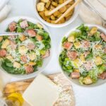 Fresh Caesar salads with grilled chicken, romaine lettuce, shredded cheese, croutons, and dressing on white plates, with a side of seasoned croutons in a bowl.