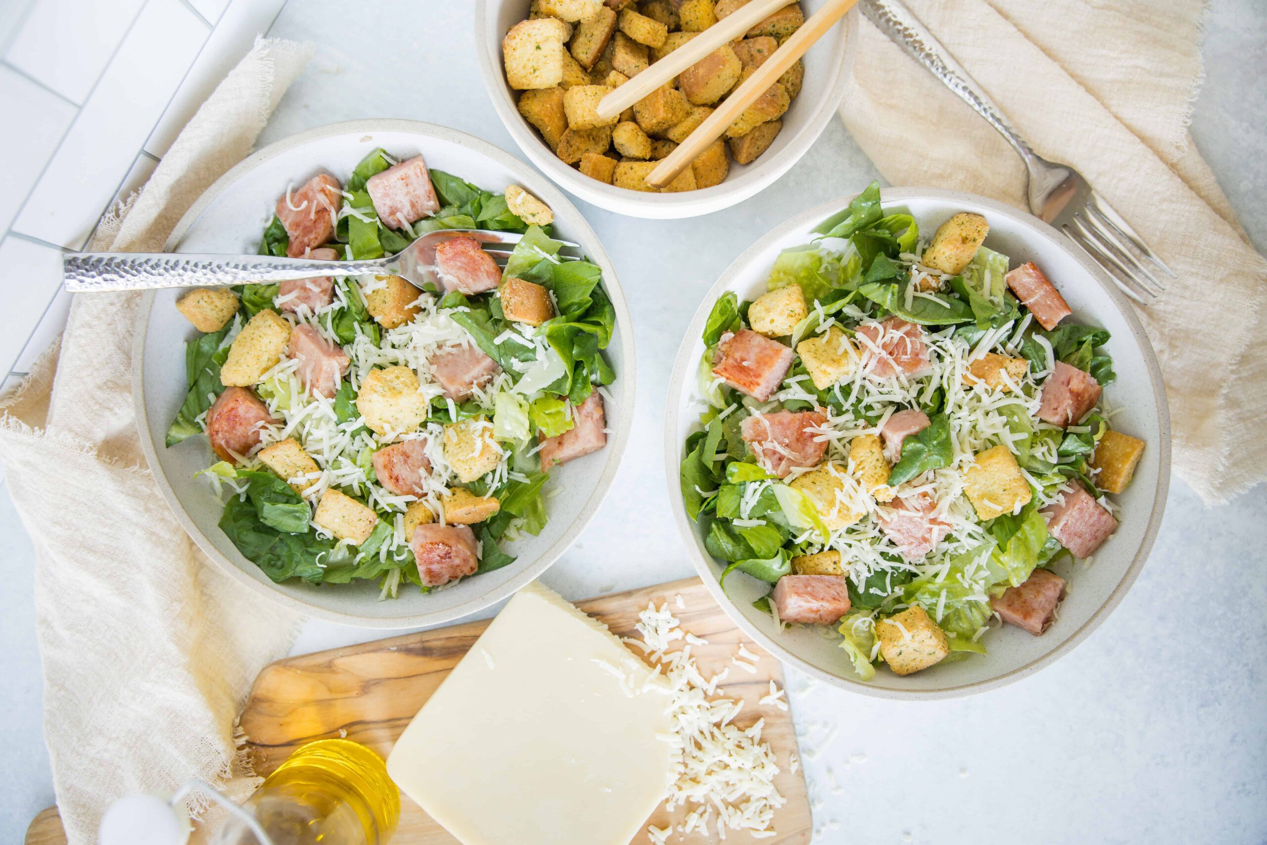Fresh Caesar salads with grilled chicken, romaine lettuce, shredded cheese, croutons, and dressing on white plates, with a side of seasoned croutons in a bowl.