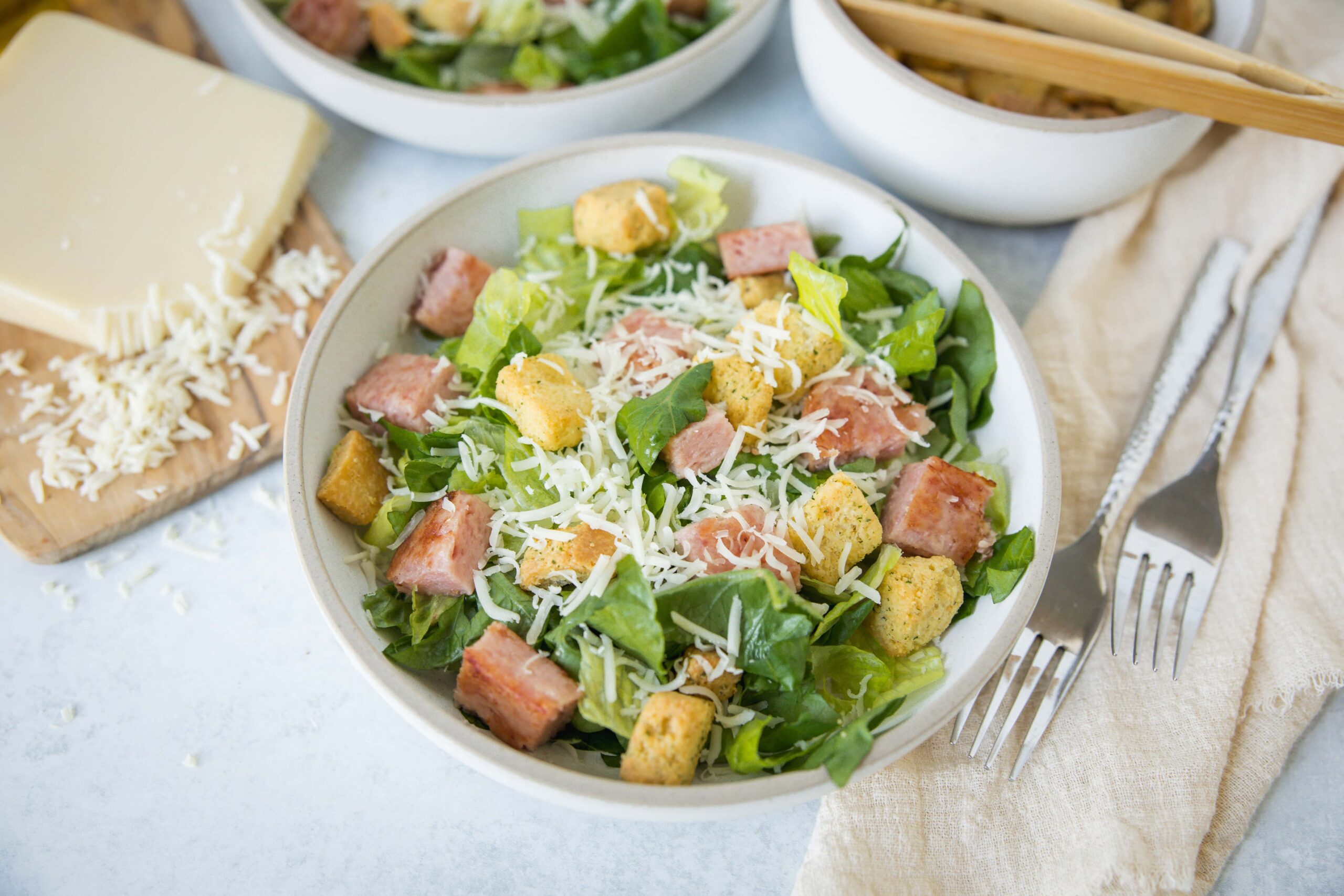 Crispy chicken Caesar salad with fresh greens, grated cheese, croutons, and bacon bits served in a white bowl on a light surface.