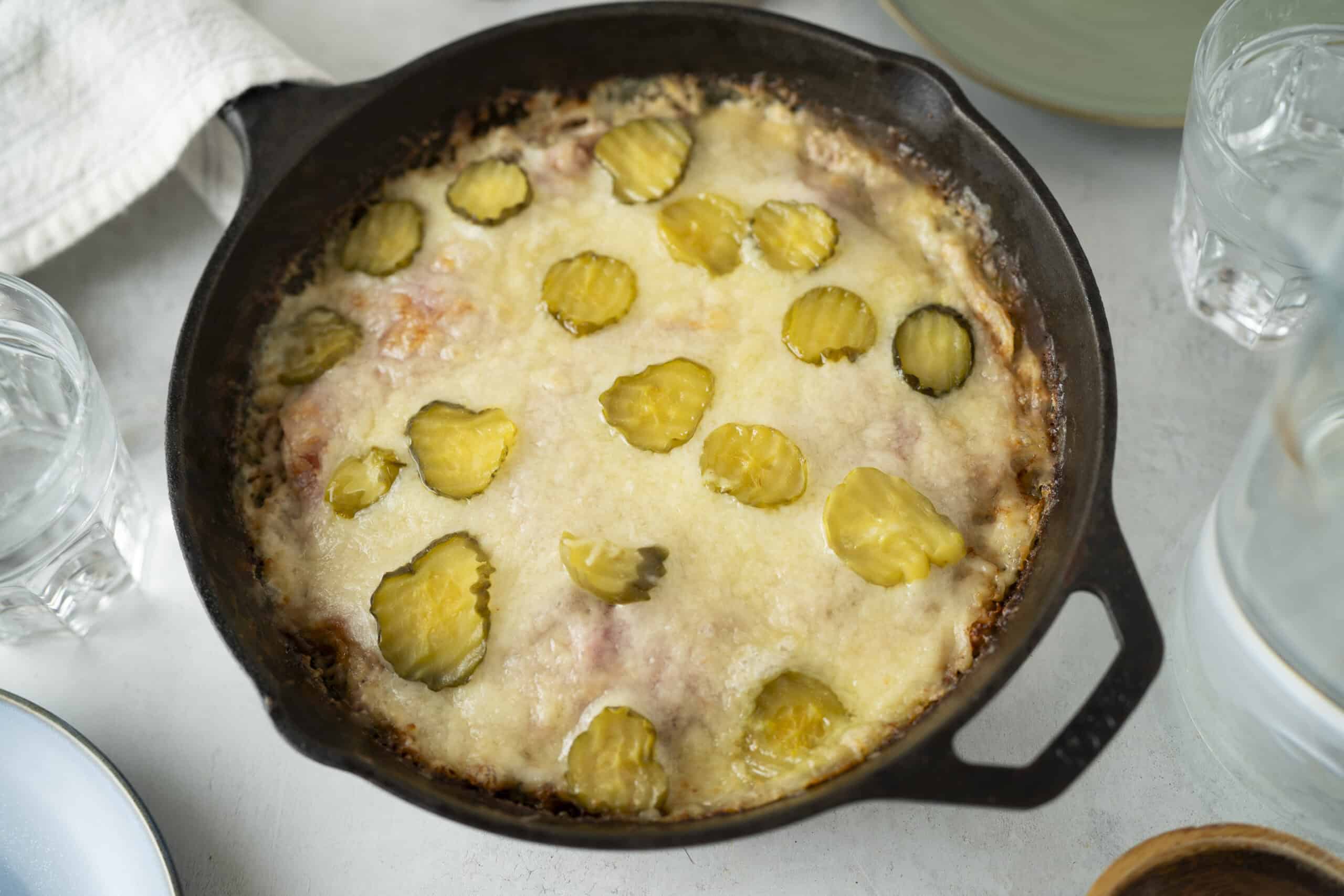 Creamy baked cheese casserole topped with sliced pickles, served in a cast iron skillet, perfect for comfort food and snack recipes.