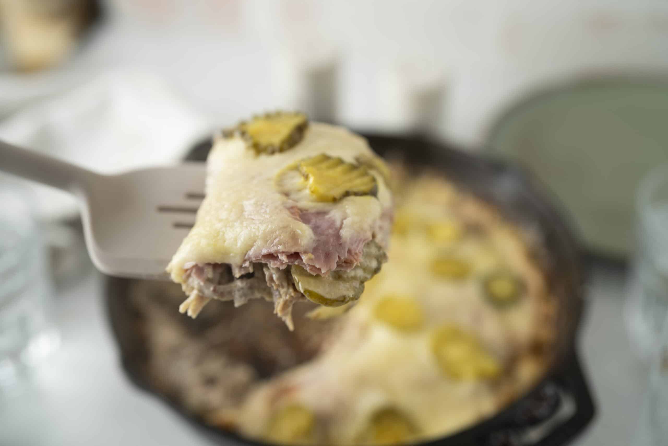 Sliced casserole dish with ham, cheese, and pickles on a spatula, ready to serve; baked casserole with melted cheese and toppings.