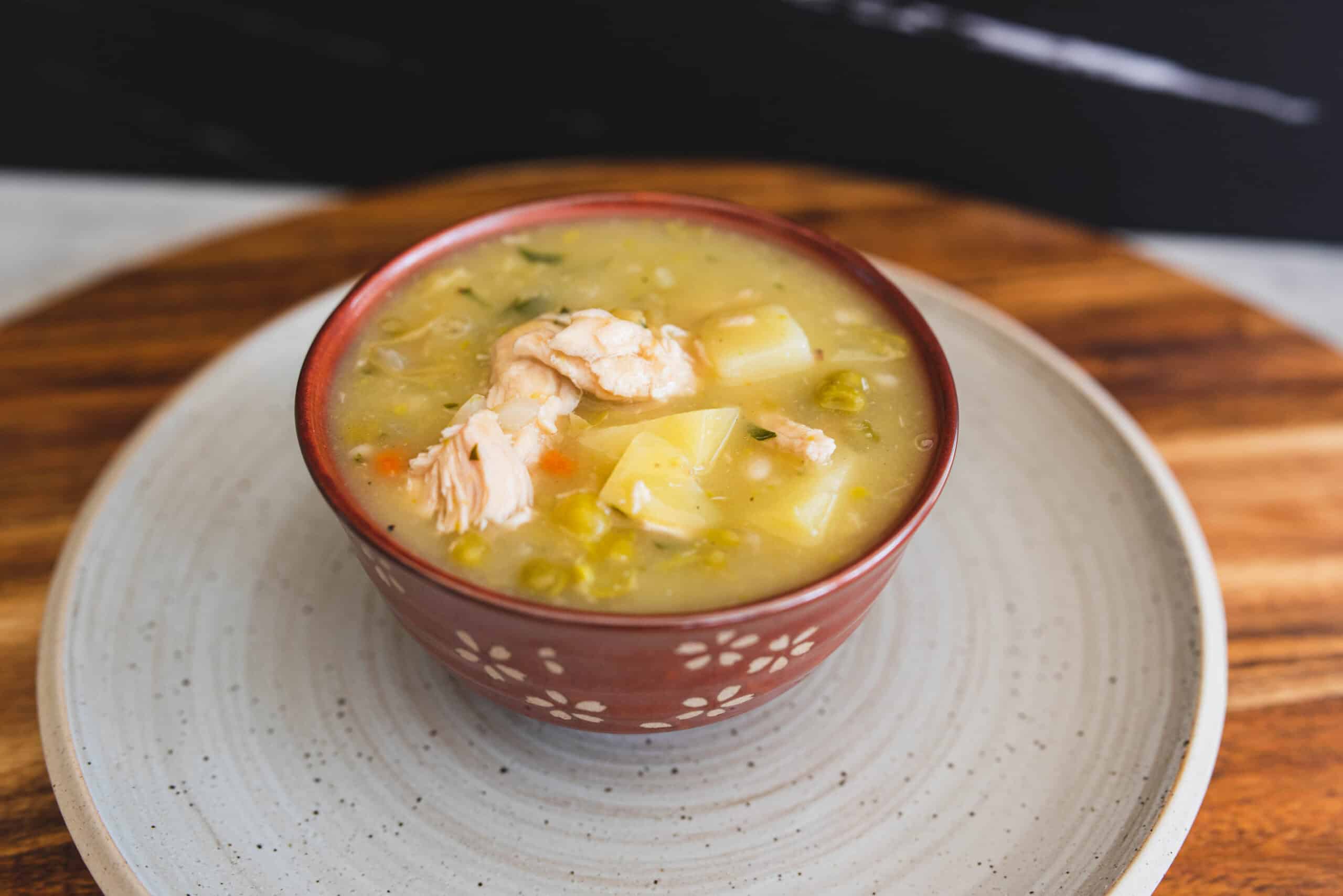 Creamy chicken and vegetable soup in a red floral-patterned bowl with potatoes, peas, and shredded chicken, served on a rustic wooden tray, perfect for comforting homemade recipes.