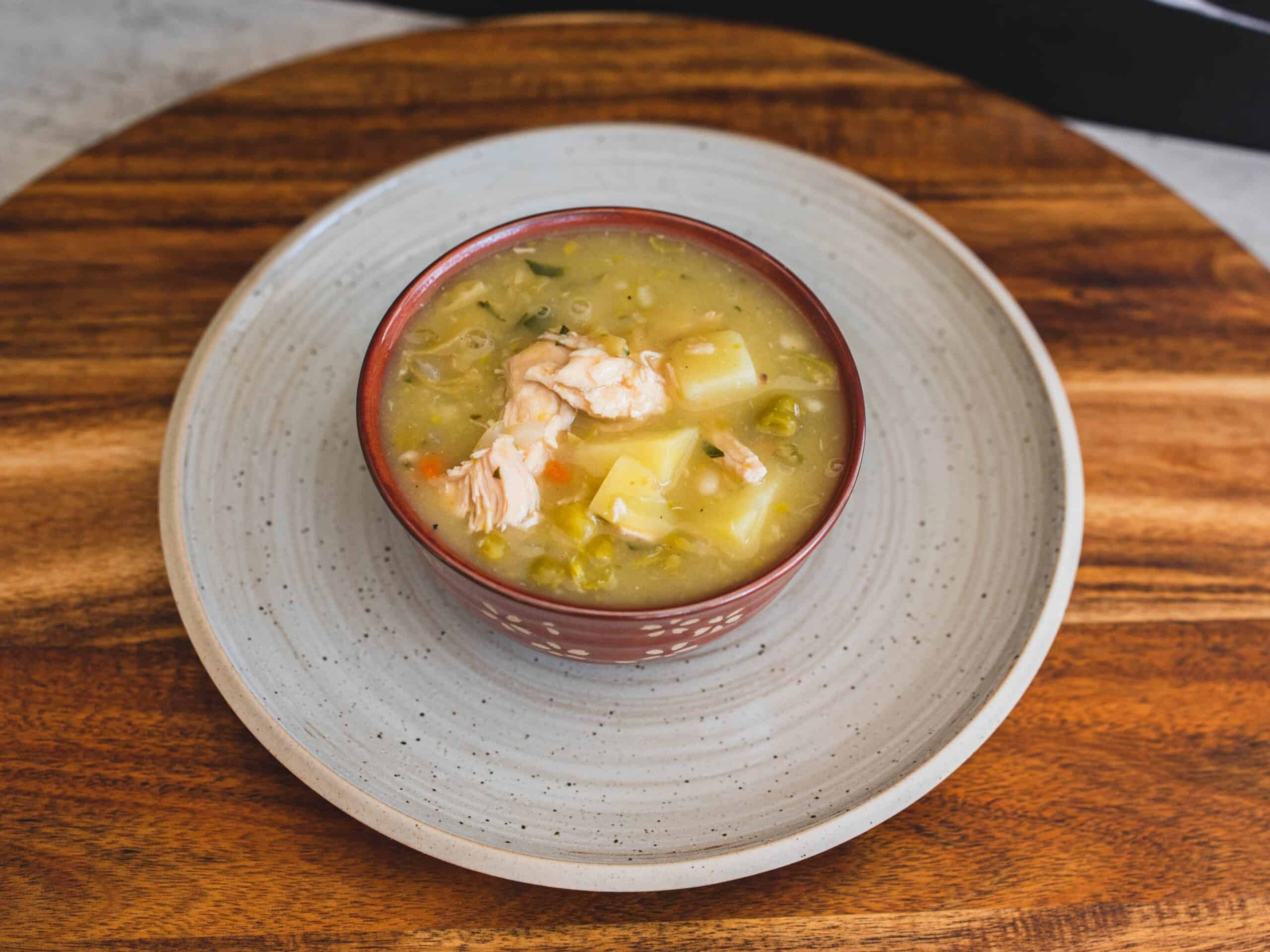 Creamy chicken and vegetable soup in a decorative bowl on a wooden tray, featuring potatoes, green peas, and shredded chicken for a hearty, nutritious meal.