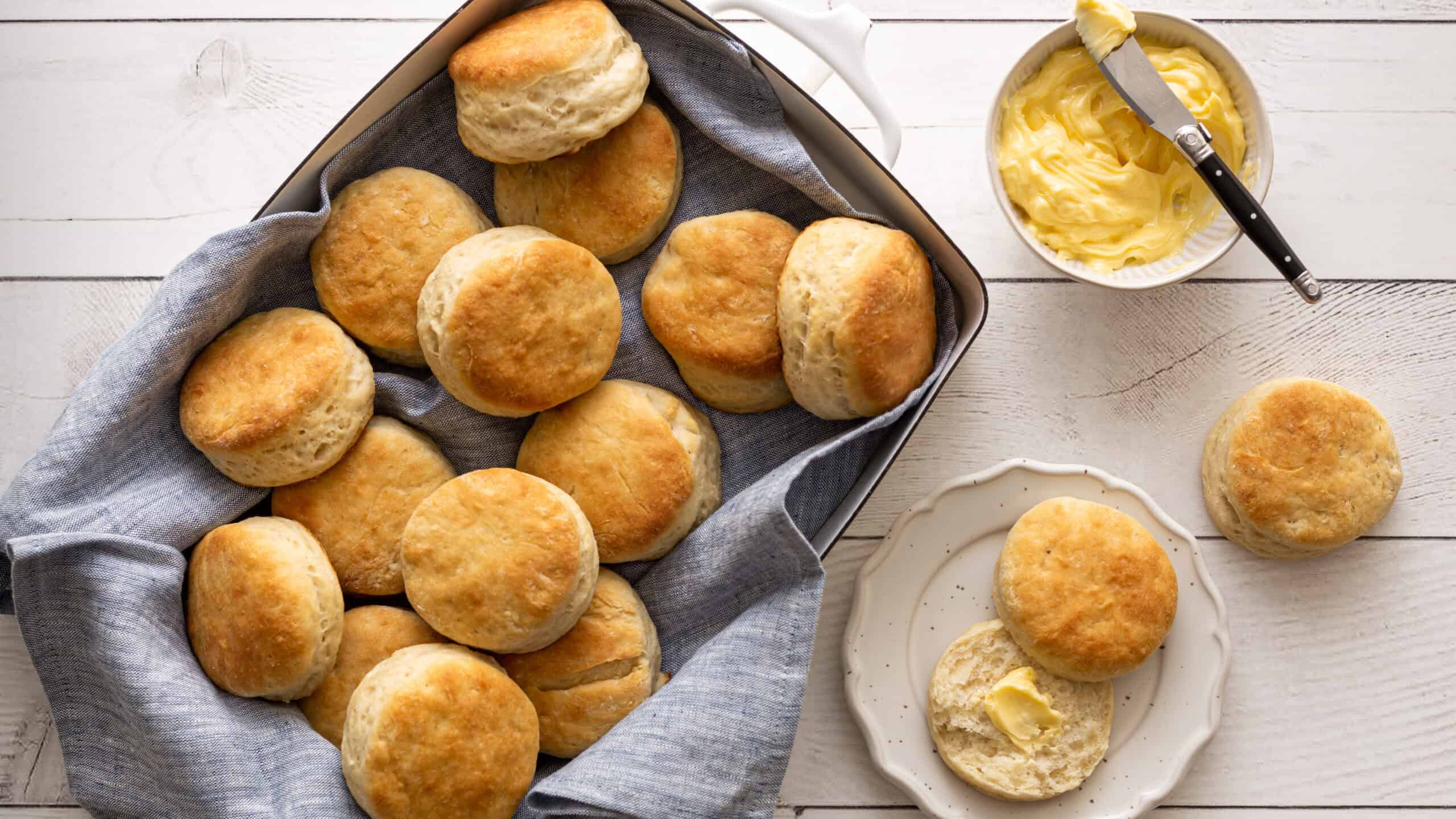 Fluffy homemade dinner rolls with a golden crust, served in a basket lined with cloth, alongside a dish of butter on a white wooden table. Freshly baked bread perfect for breakfast or dinner.