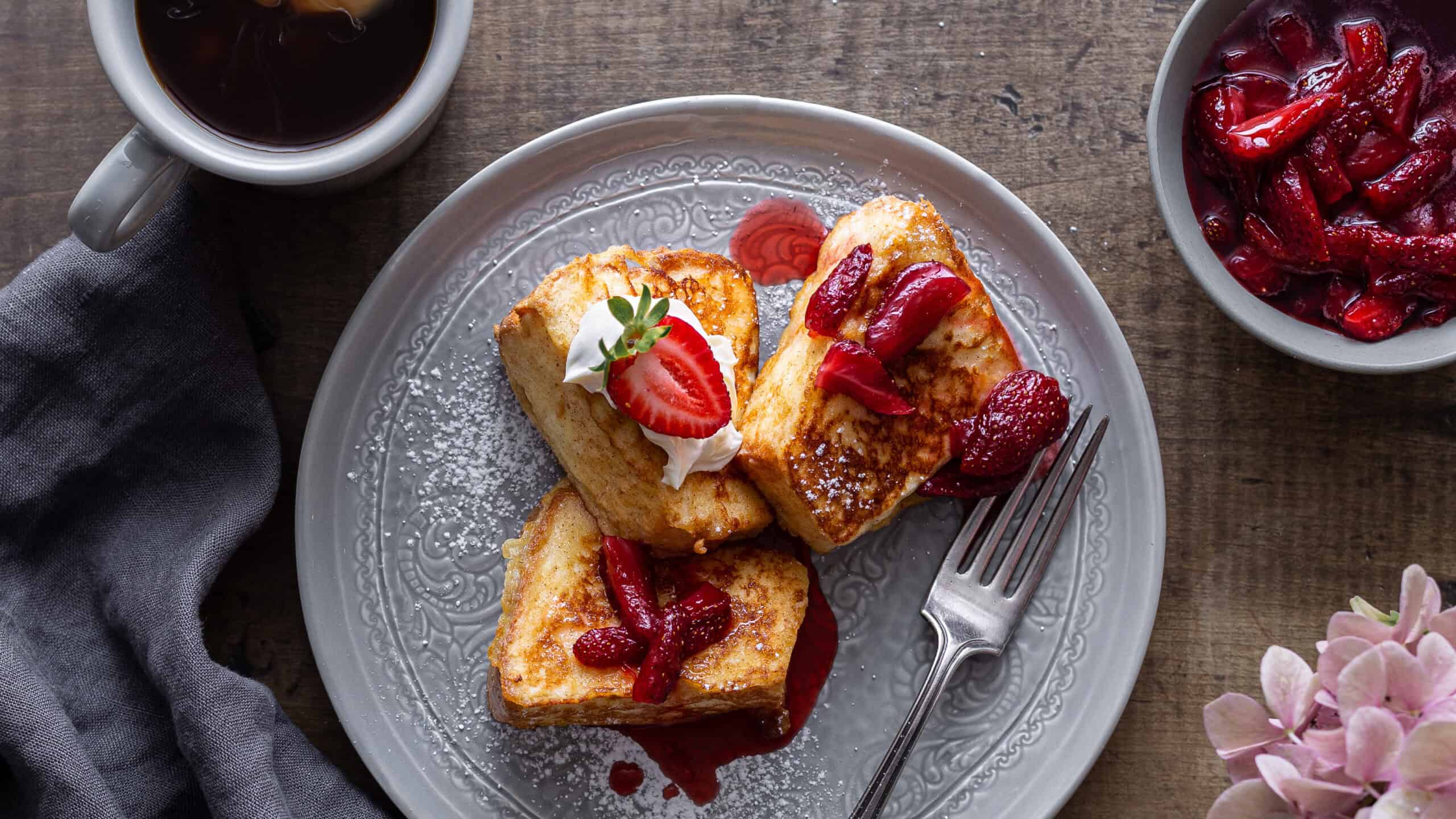 Sweet French toast topped with fresh strawberries and syrup, served on an elegant plate with a cup of coffee and side of fruit compote. Perfect for breakfast or brunch.
