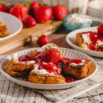 Fresh strawberry shortcake topped with whipped cream and strawberry sauce on a white plate with whole strawberries in the background.