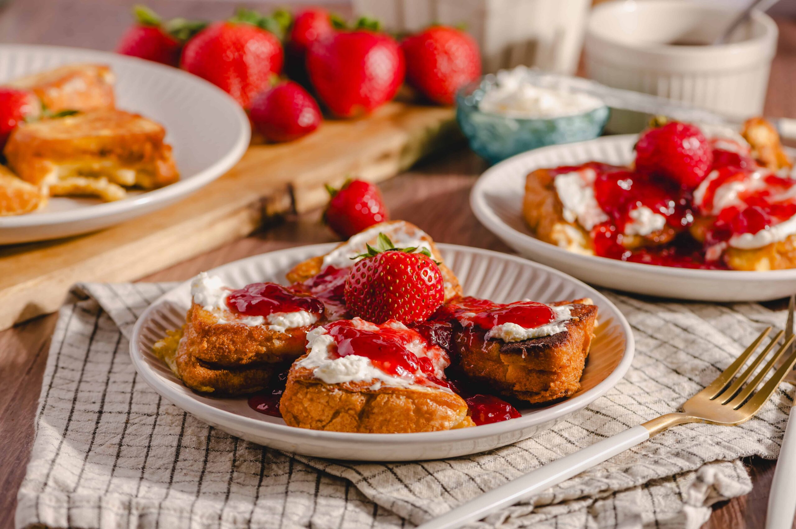 Fresh strawberry shortcake topped with whipped cream and strawberry sauce on a white plate with whole strawberries in the background.