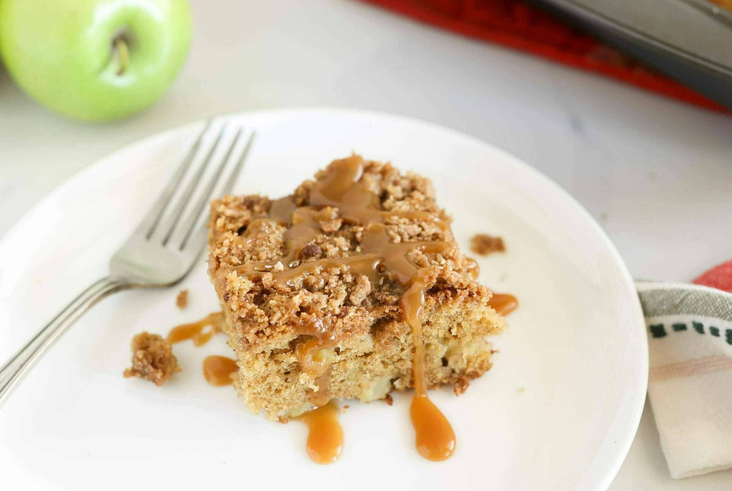 Butterscotch crumble apple cake topped with caramel drizzle, served on a white plate with a fork and green apple in the background. Perfect for fall desserts or cozy baking recipes.