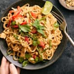 Stir-fried Thai noodle dish with vegetables, chicken, fresh herbs, and lime wedges on a black textured surface.