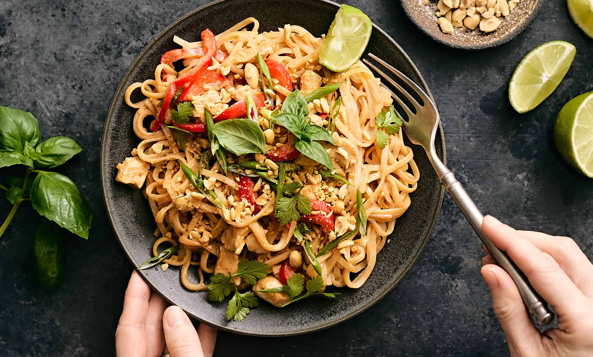 Stir-fried Thai noodle dish with vegetables, chicken, fresh herbs, and lime wedges on a black textured surface.