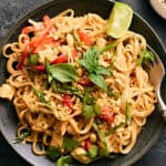 Stir-fried Thai chicken noodle dish garnished with fresh herbs, lime wedges, and chopped peanuts in a black skillet on a dark surface.