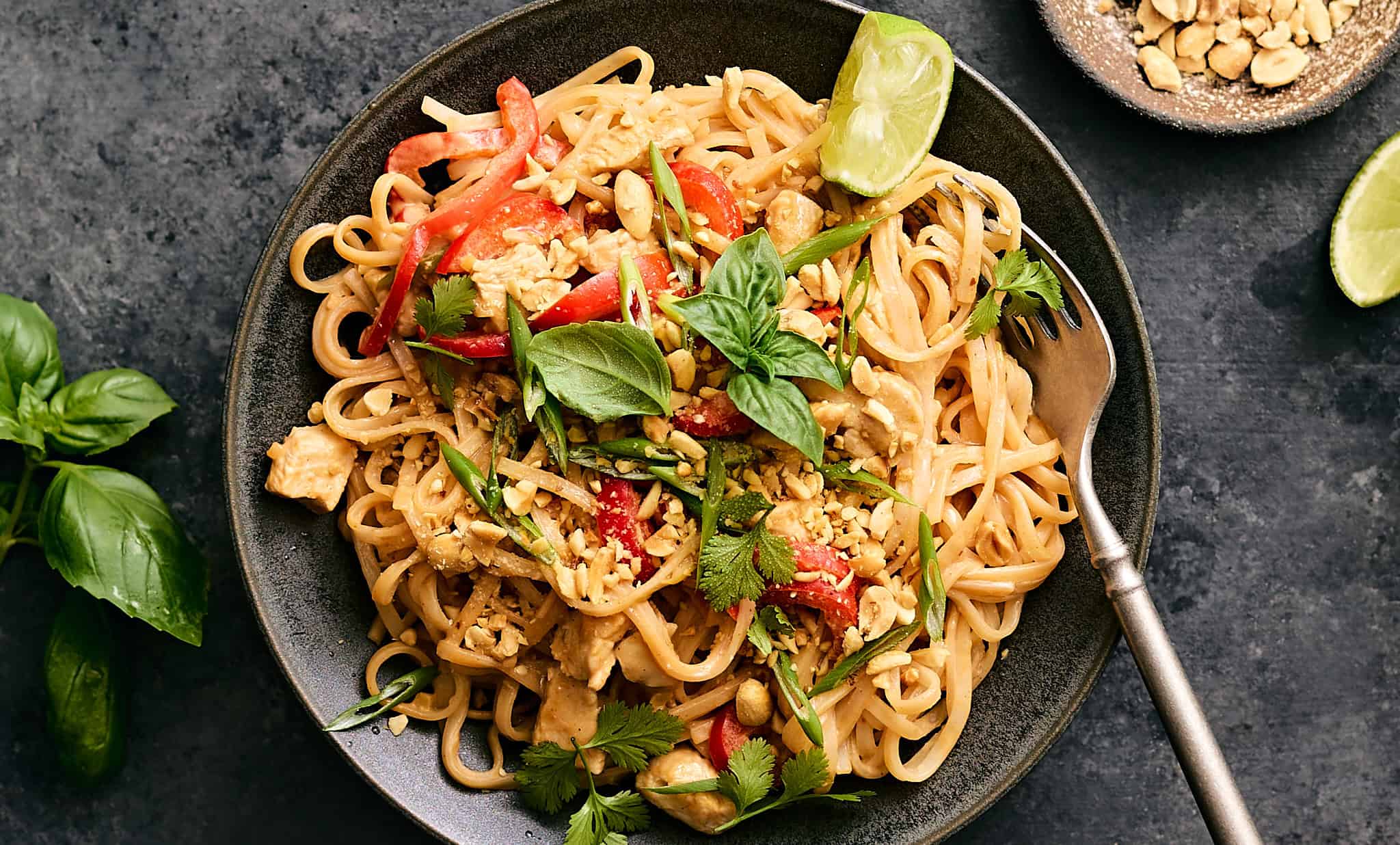 Stir-fried Thai chicken noodle dish garnished with fresh herbs, lime wedges, and chopped peanuts in a black skillet on a dark surface.