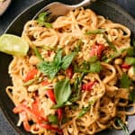 Savory Asian chicken stir-fry with noodles, fresh vegetables, crunchy peanuts, herbs, and lime wedge in black bowl on dark textured surface.