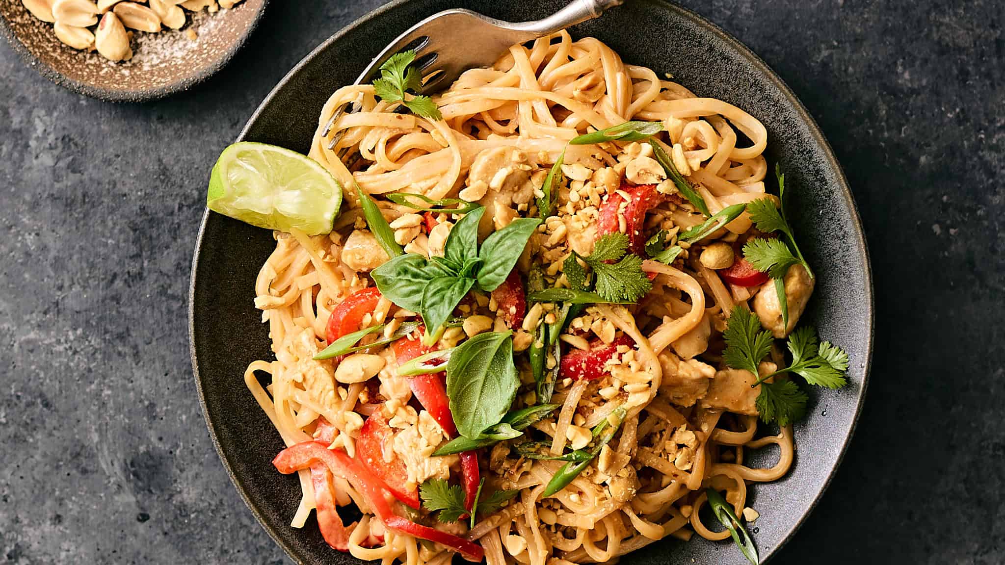 Savory Asian chicken stir-fry with noodles, fresh vegetables, crunchy peanuts, herbs, and lime wedge in black bowl on dark textured surface.