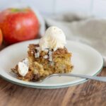 Fresh apple cinnamon crumble pie slice topped with vanilla ice cream on a white plate with whole red apples in the background.