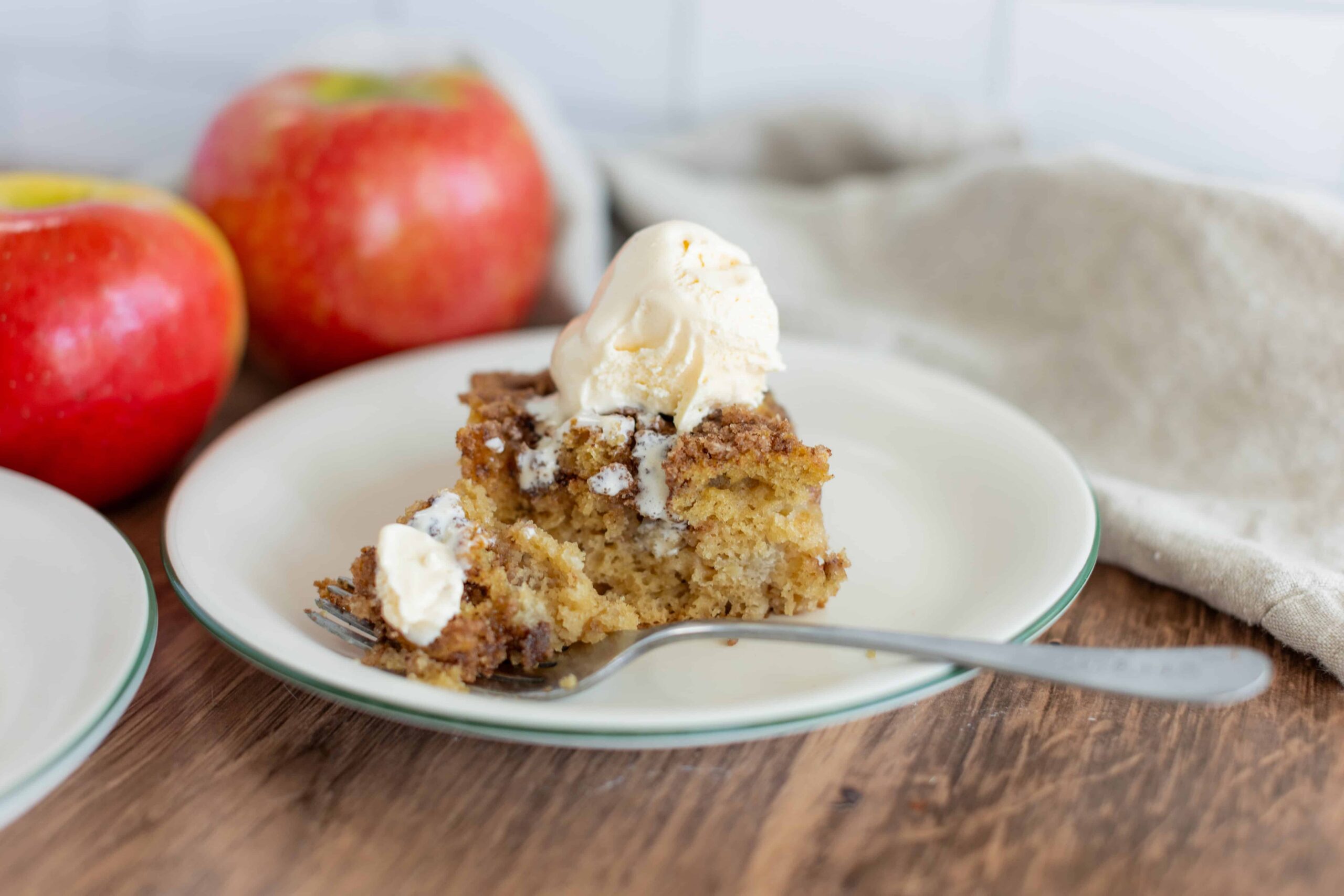 Fresh apple cinnamon crumble pie slice topped with vanilla ice cream on a white plate with whole red apples in the background.