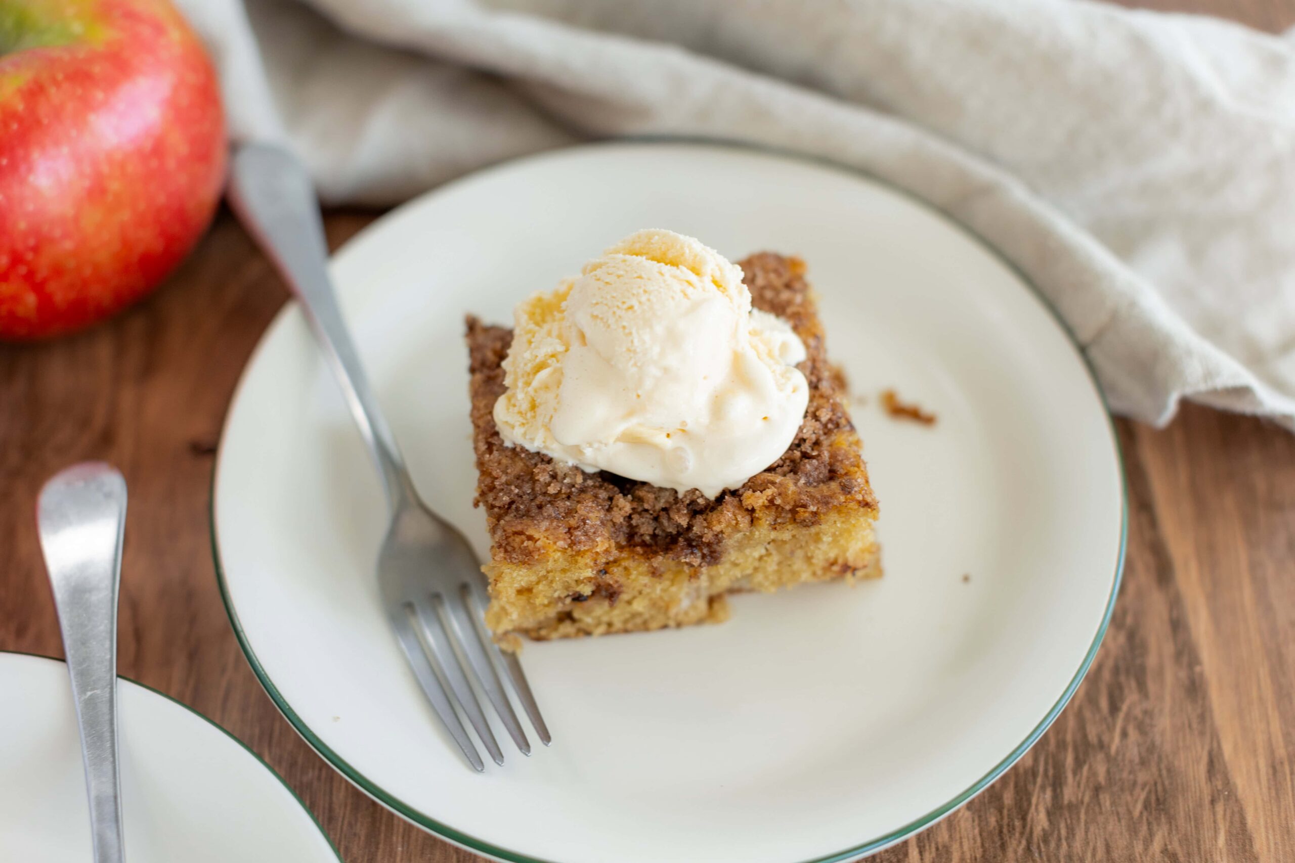 Fresh apple crumble dessert topped with vanilla ice cream on a white plate, served with a fork, with an apple and wooden table in the background.