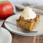 Moist apple cake topped with vanilla ice cream on a white plate, with fresh apples in the background. Perfect for apple dessert recipes and fall baking.