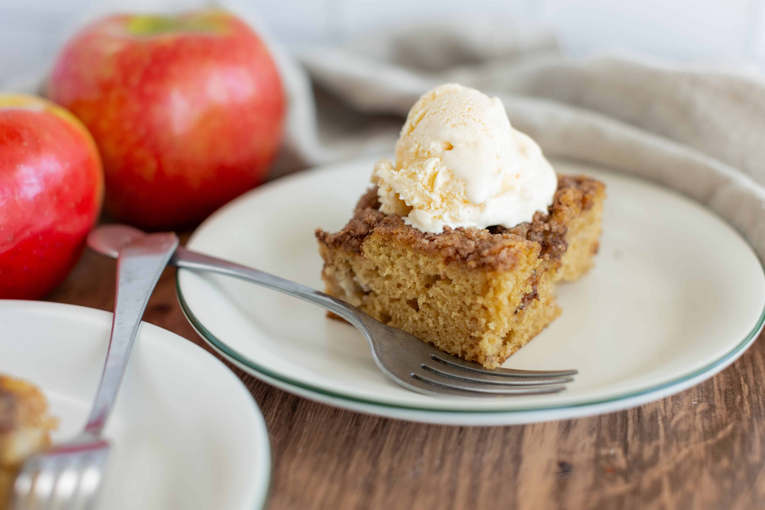 Moist apple cake topped with vanilla ice cream on a white plate, with fresh apples in the background. Perfect for apple dessert recipes and fall baking.