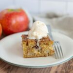 Fresh apple cinnamon cake slice topped with vanilla ice cream on a white plate, with whole red apples in the background, perfect for apple dessert recipes and fall baking ideas.