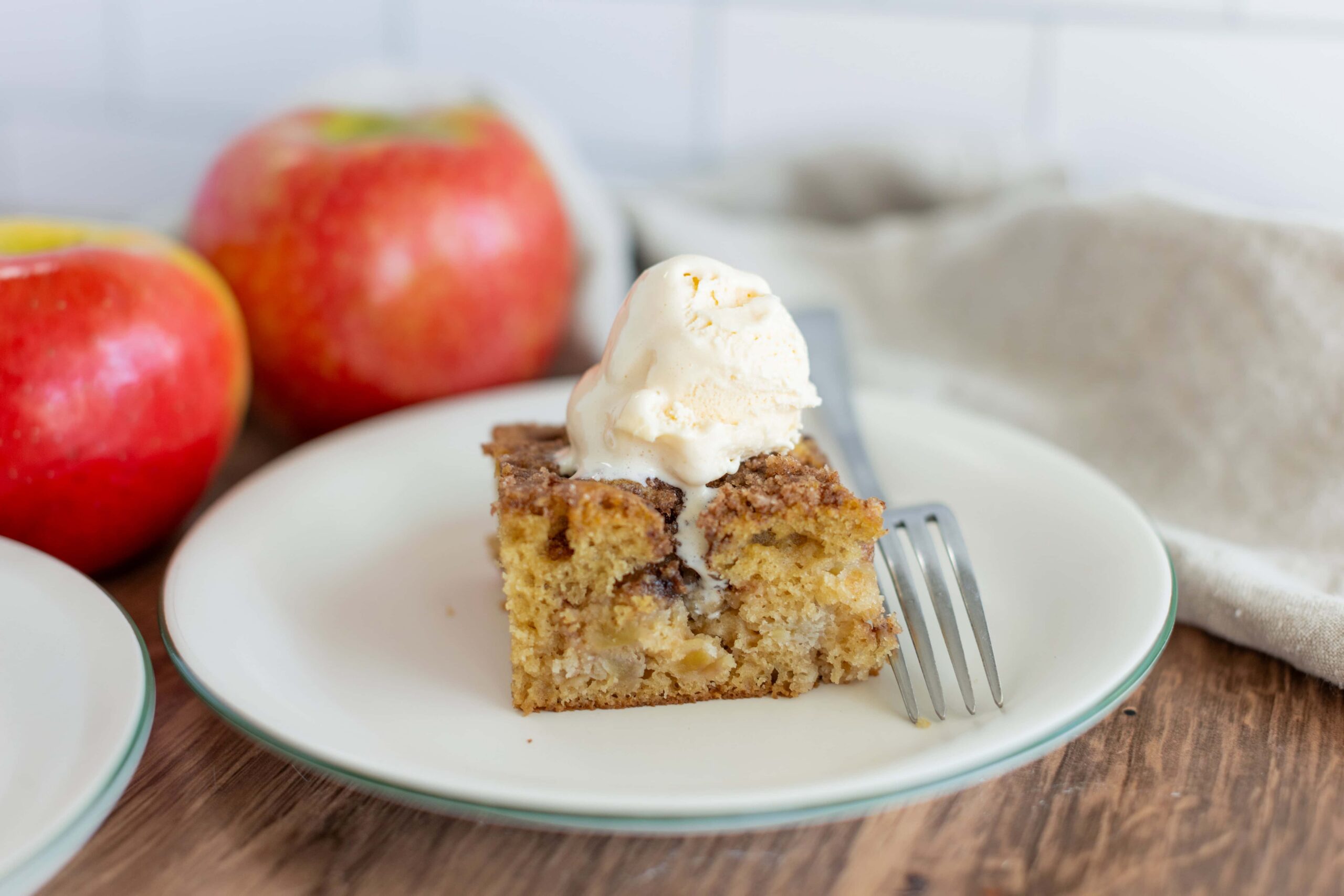 Fresh apple cinnamon cake slice topped with vanilla ice cream on a white plate, with whole red apples in the background, perfect for apple dessert recipes and fall baking ideas.