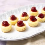Mini cheesecakes topped with cherries, served on a white decorative platter. Perfect for desserts, parties, or special occasions. Deliciously creamy with a cherry garnish.