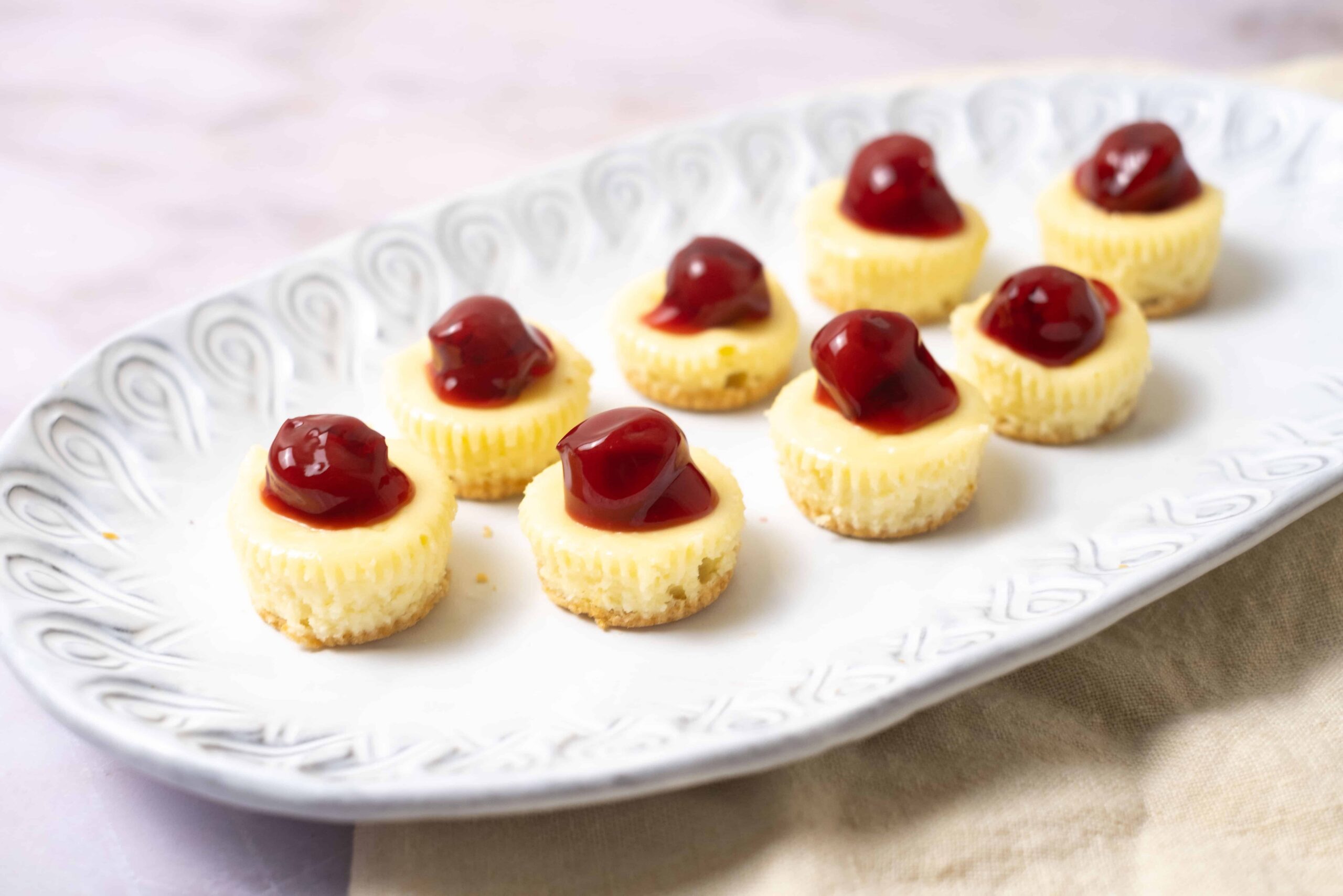 Mini cheesecakes topped with cherries, served on a white decorative platter. Perfect for desserts, parties, or special occasions. Deliciously creamy with a cherry garnish.