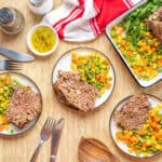 Juicy meatloaf with mixed vegetables on plates, baked meatloaf with fresh green parsley garnish, served with colorful steamed mixed vegetables, placed on a wooden table with cutlery and seasonings, appetizing home-cooked dinner.
