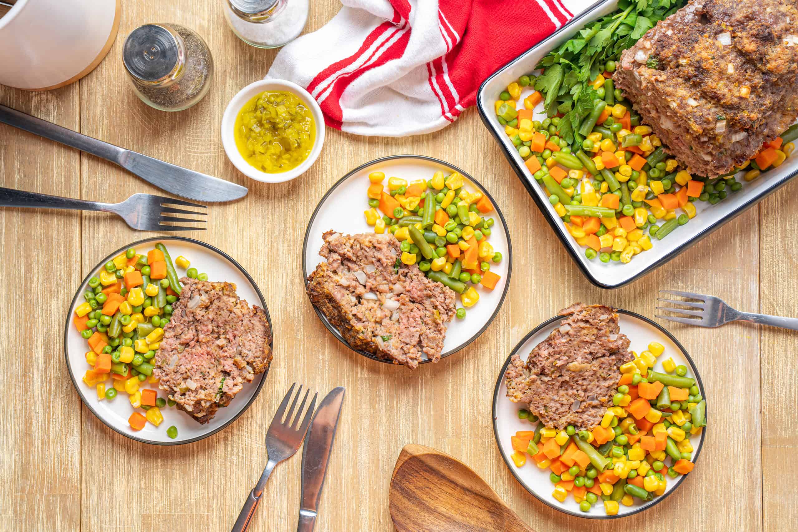 Juicy meatloaf with mixed vegetables on plates, baked meatloaf with fresh green parsley garnish, served with colorful steamed mixed vegetables, placed on a wooden table with cutlery and seasonings, appetizing home-cooked dinner.