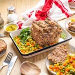 Juicy meatloaf with mixed vegetables on a wooden table, topped with fresh herbs, and served with mashed carrots, green beans, corn, and peas. Prepared for a hearty family dinner or special occasion.