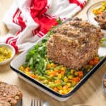 Ground meat meatloaf with mixed vegetables and fresh herbs on a white baking dish, served with side dishes of cooked peas, carrots, corn, and green beans for a hearty, homemade dinner.