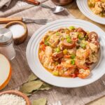 Savory seafood and sausage paella served on a white plate with garnishes, accompanied by ingredients like rice, onion, and spices, on a rustic wooden table.