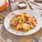 Sausage, chicken, shrimp, and vegetable stir-fry on a white plate with fresh green onions, served on a rustic table with ingredients like rice, salt, pepper, bay leaves, and spices.