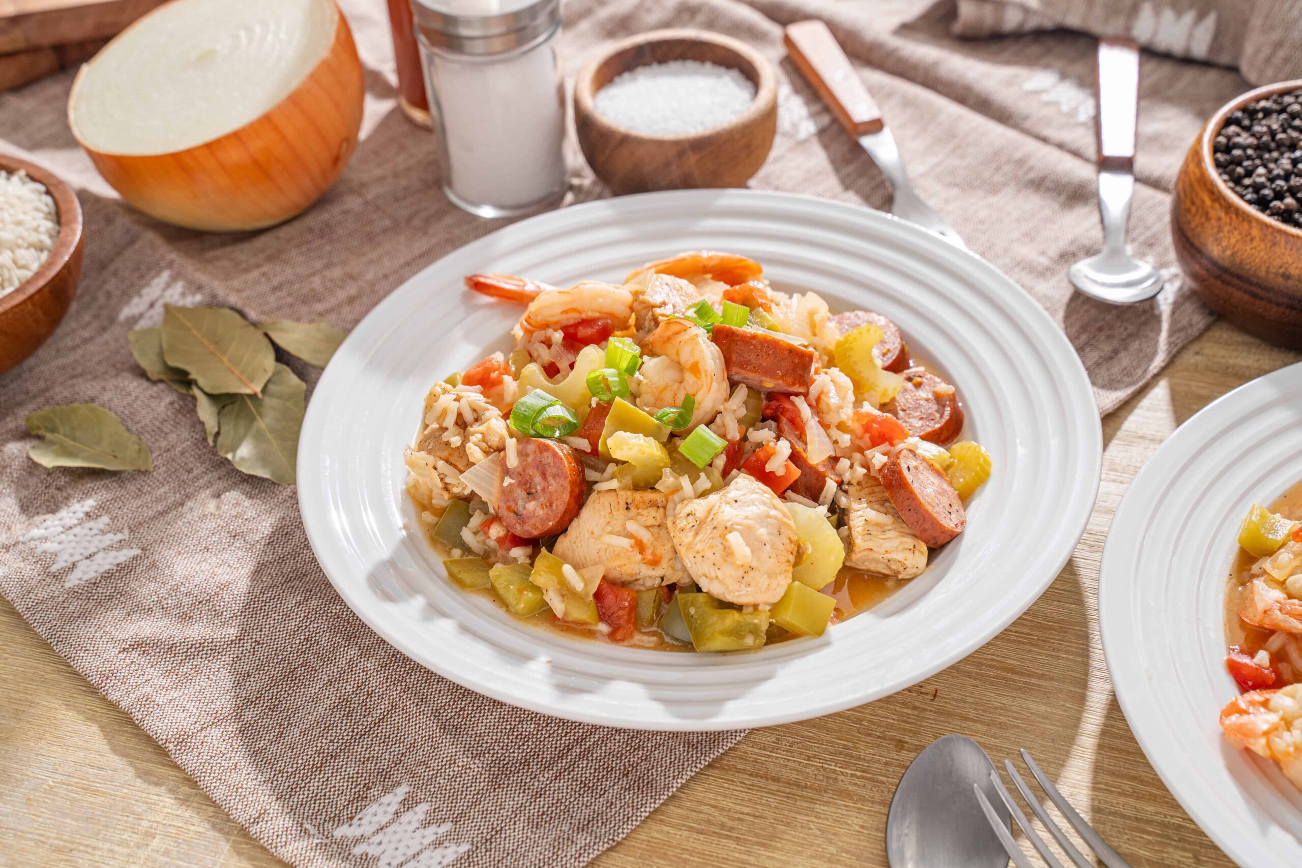 Sausage, chicken, shrimp, and vegetable stir-fry on a white plate with fresh green onions, served on a rustic table with ingredients like rice, salt, pepper, bay leaves, and spices.