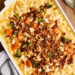 Cheesy casserole with vegetables and crispy bacon topping, served in a white baking dish on a wooden surface, garnished with fresh salad greens on the side.