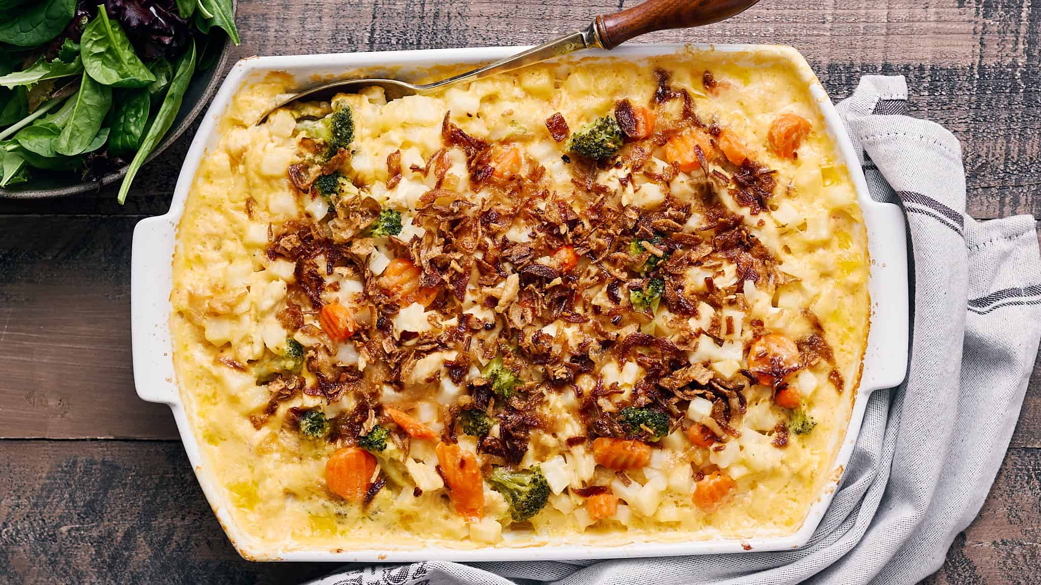 Creamy vegetable and chicken casserole topped with crispy fried onions in a white baking dish on rustic wooden table, garnished with fresh greens.