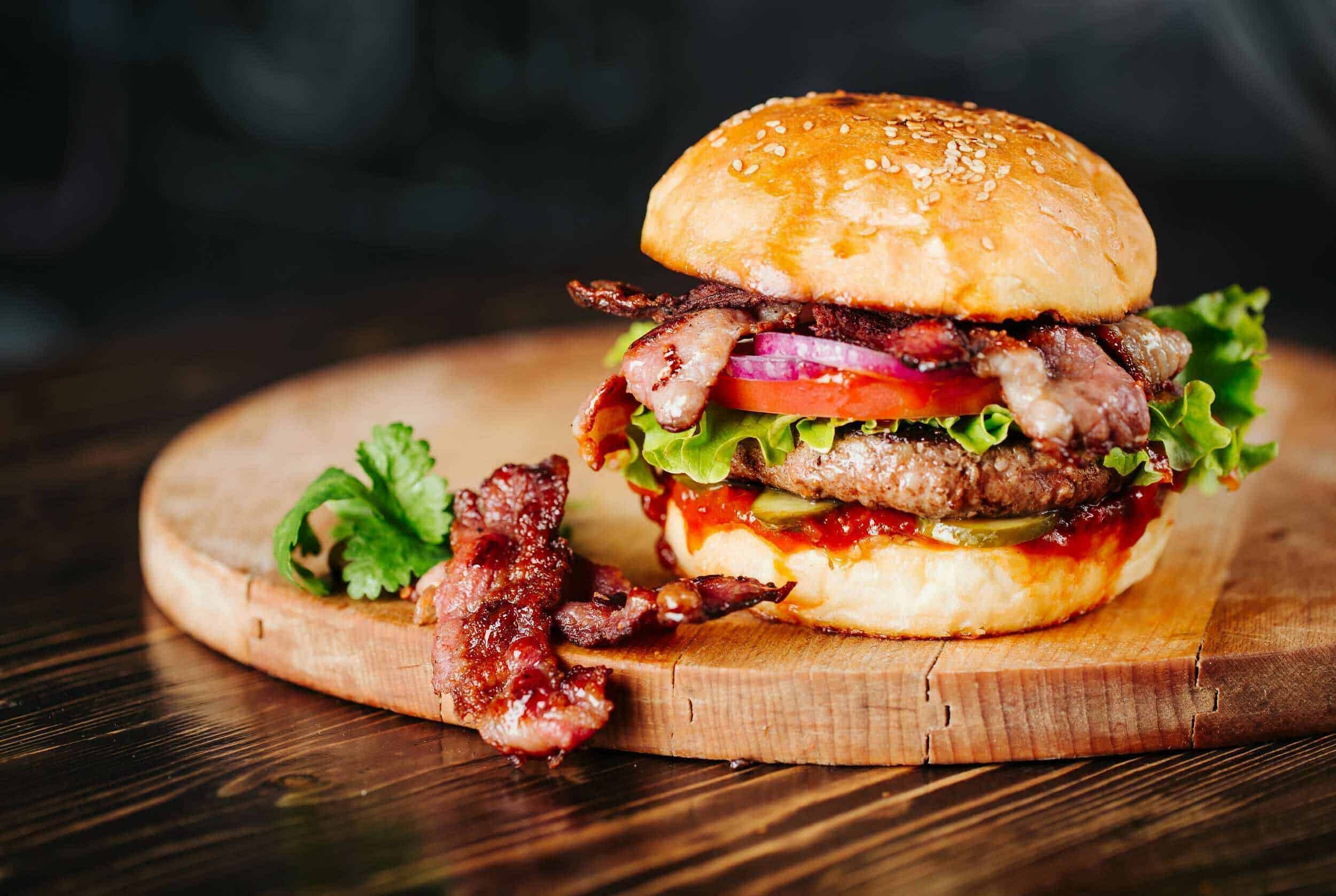Juicy bacon cheeseburger with fresh vegetables on a wooden board, featuring lettuce, tomato, onion, and pickles, perfect for classic American comfort food.